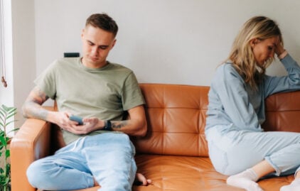 Ein junges Paar sitzt auf einer Couch. Während der Mann am Handy ist, sitzt die Frau abgewandt ein Stück neben ihm. Dei Stimmung wirkt angespannt.